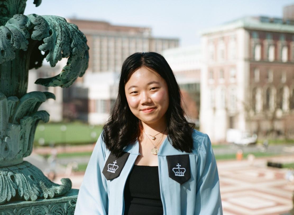 Young Asian woman, smiling with her mouth closed, dressed in Columbia University graduation gown. The background is the University