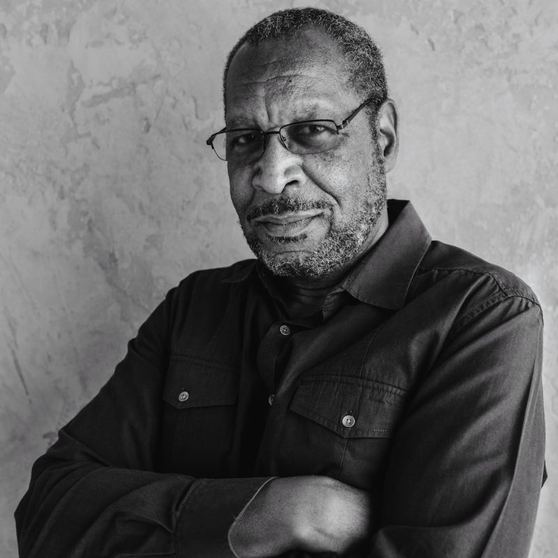 Older Black man with wire framed glasses, a scruffy beard, and his arms crossed across his chest