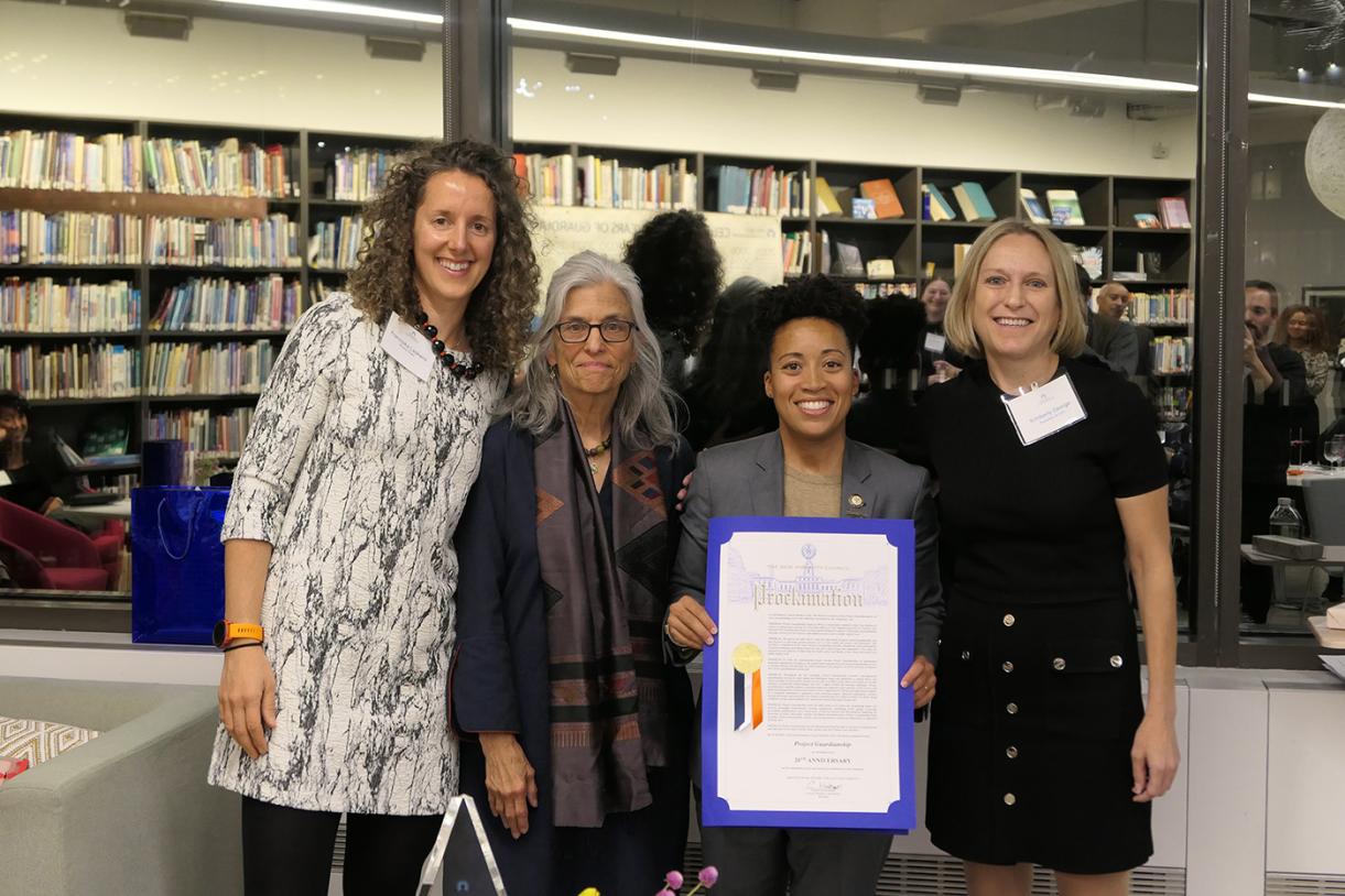 Four women of various heights holding a large sheet of paper with blue trimming.