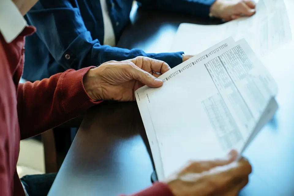 a person holding a budget sheet. the person is wearing a burgundy shirt and they are sitting next to someone wearing a blue shirt.
