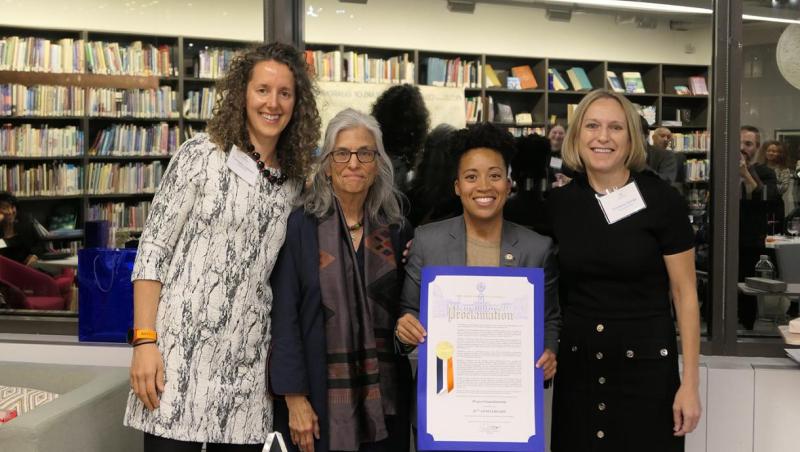 Four women of various heights holding a large sheet of paper with blue trimming.