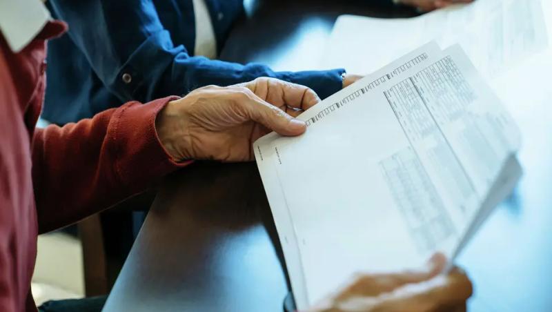 a person holding a budget sheet. the person is wearing a burgundy shirt and they are sitting next to someone wearing a blue shirt.