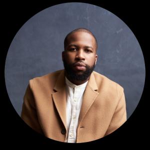 A bald male with a beard. He is wearing a camel jacket over a white shirt.